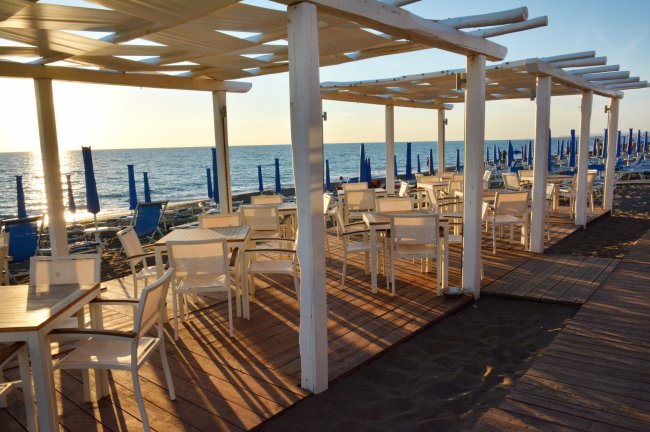 Tavoli e sedie sotto una pergola bianca sulla spiaggia, vista mare al tramonto.