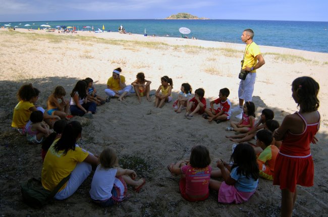 Gruppo di bambini seduti in cerchio sulla sabbia con un adulto. Spiaggia e mare sullo sfondo.
