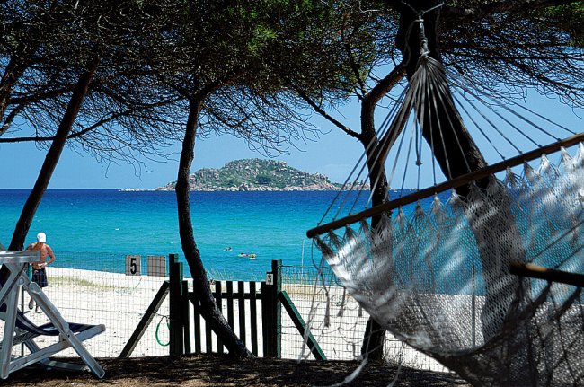 Amaca su una spiaggia sabbiosa con mare turchese e isola in lontananza, vista tra gli alberi.