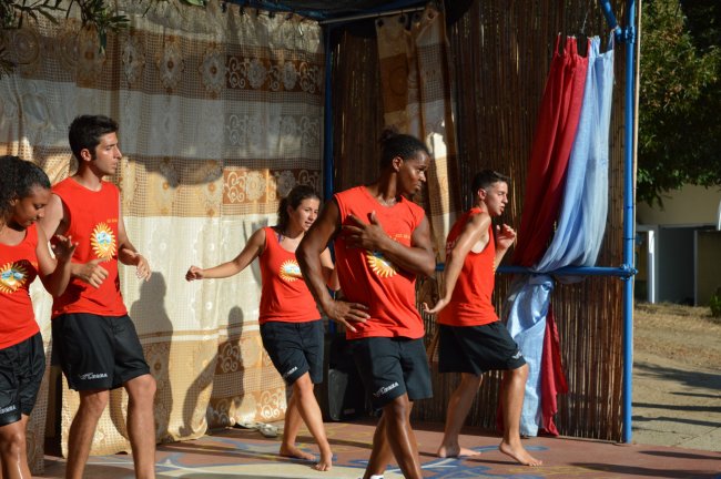 Gruppo di giovani danza su un palco. Indossano canotte rosse e pantaloncini neri.