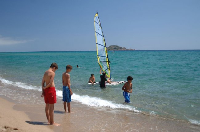 Ragazzi sulla spiaggia osservano un windsurfista nel mare blu con piccola isola sullo sfondo.