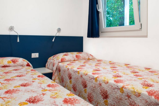 Due letti singoli con copriletto a motivi di coralli colorati, accanto a una finestra con tende blu.