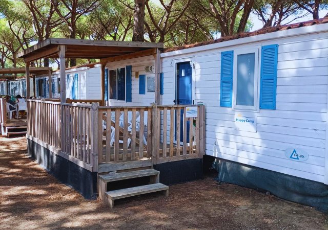 Esterno di un bungalow bianco con persiane blu e veranda in legno, situato in un'area boschiva.