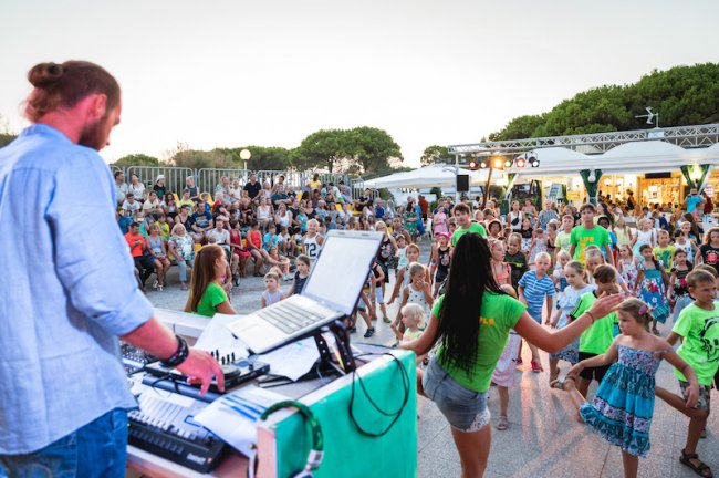 DJ alla console mentre bambini e istruttori ballano, pubblico dietro.