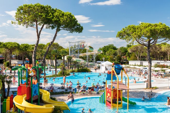 Parco acquatico con piscine azzurre, scivoli gialli e alberi alti.