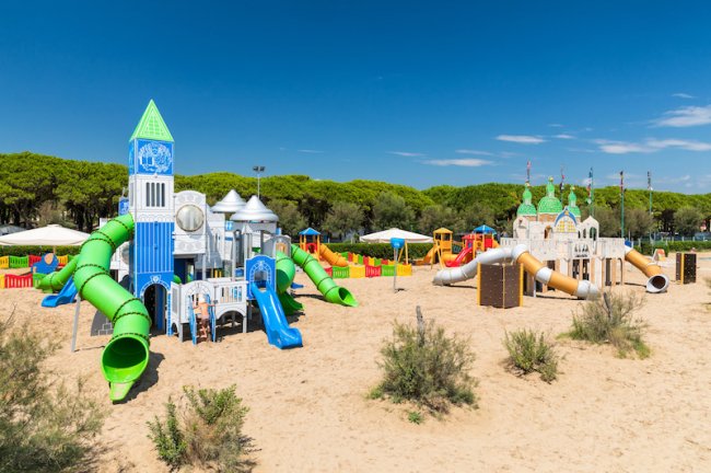 Parco giochi sabbioso con scivoli verdi e blu, torri e tema di castello sotto un cielo azzurro.