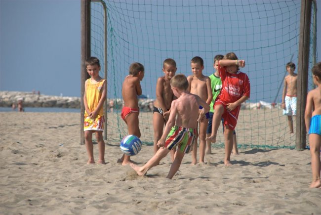 Bambini giocano a calcio in spiaggia, un bambino calcia la palla verso una piccola porta.