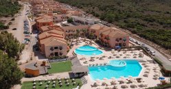 Complesso con due piscine, ombrelloni beige, edifici color pesca e vegetazione circostante.