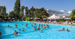 Persone in una piscina all'aperto, circondata da alberi e ombrelloni.