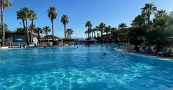 Ampia piscina con palme, persone che nuotano, cielo blu.