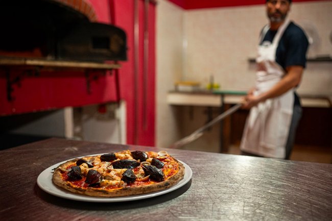 Pizza su un tavolo, forno a legna e pizzaiolo sullo sfondo.