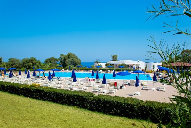 Piscina con lettini e ombrelloni blu, mare sullo sfondo.