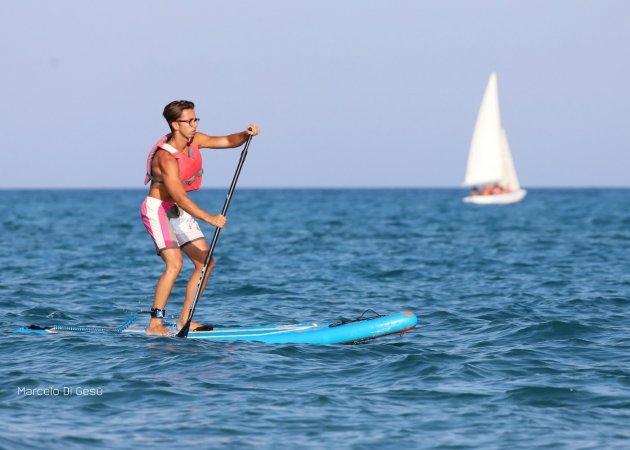 Uomo su tavola da paddle in mare con una barca a vela.