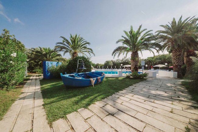 Una barca blu posizionata su un prato fra palme, con una piscina visibile sullo sfondo.