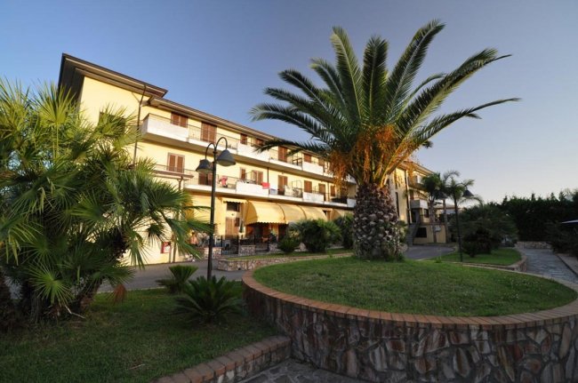 Edificio a tre piani con palme e giardino curato davanti.