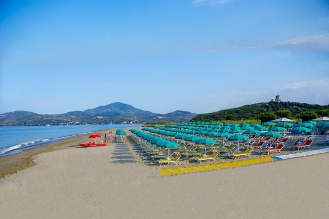 Spiaggia con lettini, ombrelloni turchesi, pedalò rossi. Montagne sullo sfondo e mare calmo.