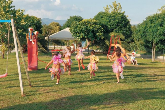 Bambini vestiti da fate corrono in un parco giochi con uno scivolo rosso, circondati da alberi.