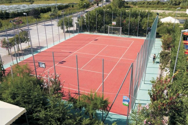 Un campo sportivo con superficie rossa, circondato da recinzioni e vegetazione.