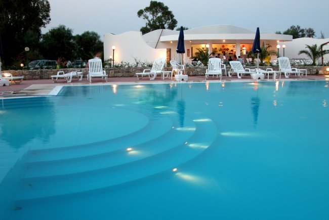 Piscina con gradini illuminati di sera, lettini bianchi e ombrelloni blu.