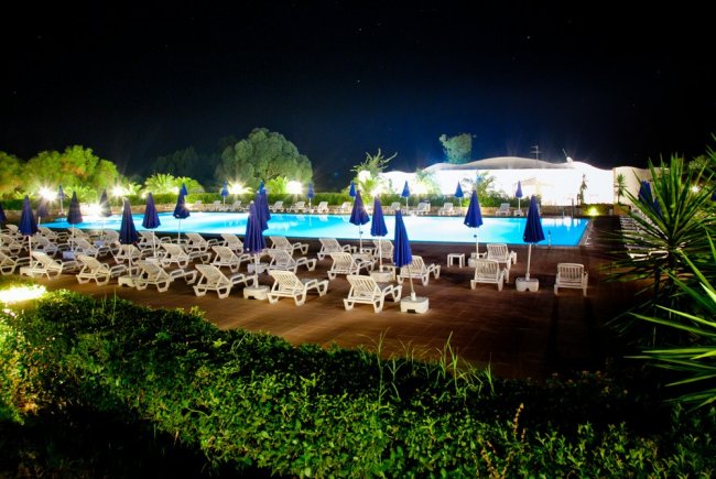 Piscina illuminata di notte, contornata da lettini e ombrelloni blu.