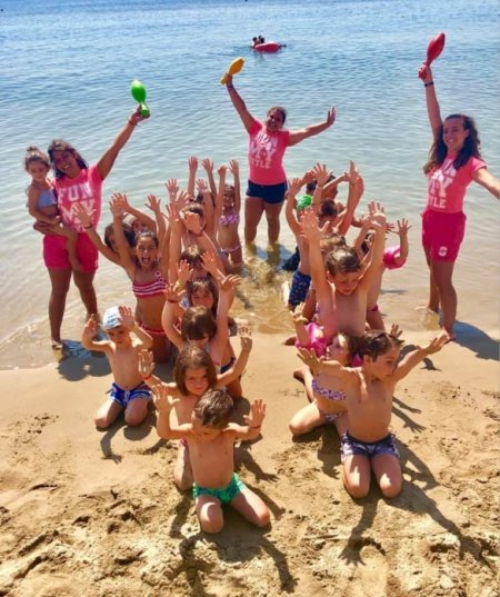 Bambini giocano in spiaggia con animatori che alzano le braccia e tengono palette colorate.