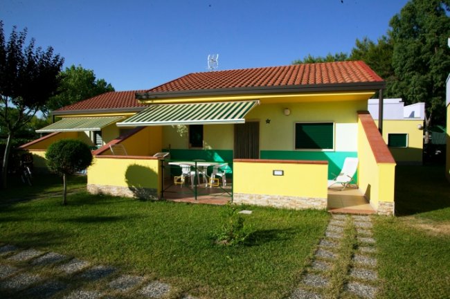 Un bungalow giallo con tetto rosso, giardino e veranda con tavolo.