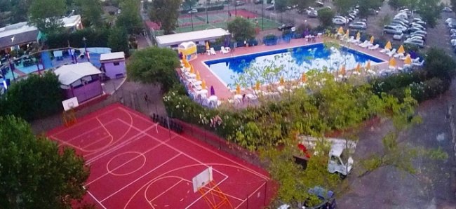 Un campo da basket rosso e una grande piscina con lettini e ombrelloni gialli.