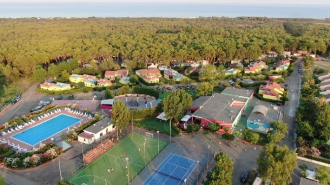 Vista aerea di una struttura turistica con piscina, campi sportivi e case tra gli alberi.
